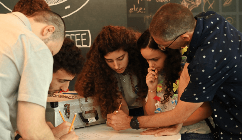  Un groupe de quatre personnes est réuni autour d'une table blanche, concentré sur la rédaction et la discussion de documents. Parmi leurs tâches, l'homme le plus âgé intègre les principes de détection et de réponse gérées tout en aidant deux personnes plus jeunes dans leur travail.