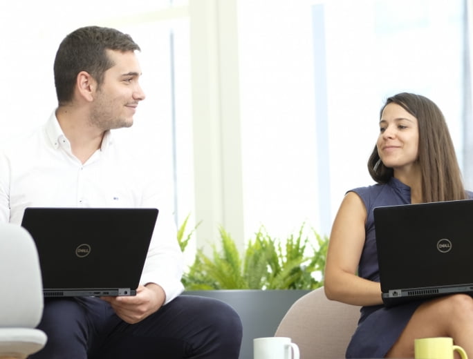  Two people are sitting indoors, smiling at each other, each using a Dell laptop. There are plants and large windows in the background, offering a serene environment as they discuss their Managed Detection and Response strategies.