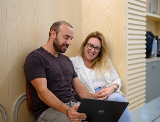  Two people sit next to a wooden wall, looking at a laptop. The man in a dark shirt and the woman in a white jacket and glasses appear engaged and smiling, possibly discussing the latest in Adaptive Managed xDR solutions.