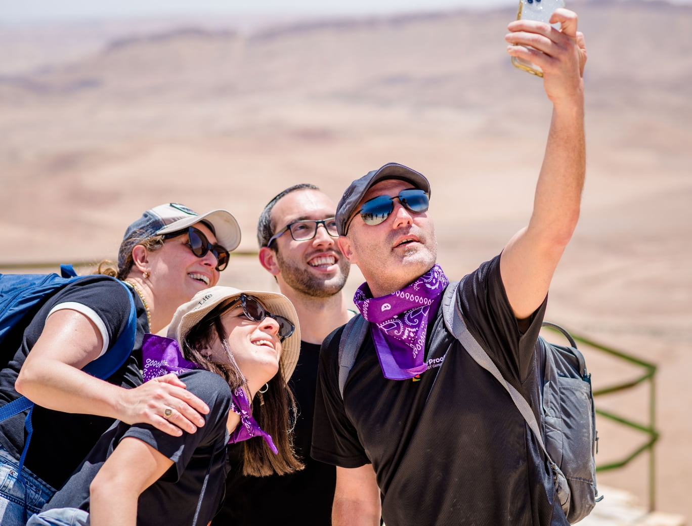  Four people taking a selfie outdoors, wearing casual clothing, sunglasses, and hats with a desert landscape in the background—a perfect snapshot of fun moments amidst the sand dunes, much like Adaptive Managed xDR providing security amidst the digital wilderness.