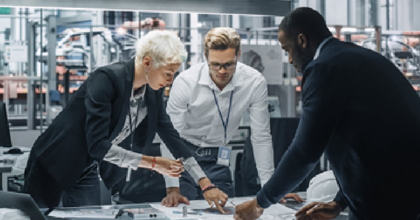 Three people in business attire collaborate on a project, examining documents on a table in a modern industrial setting, strategizing their next steps using Adaptive Managed xDR solutions.