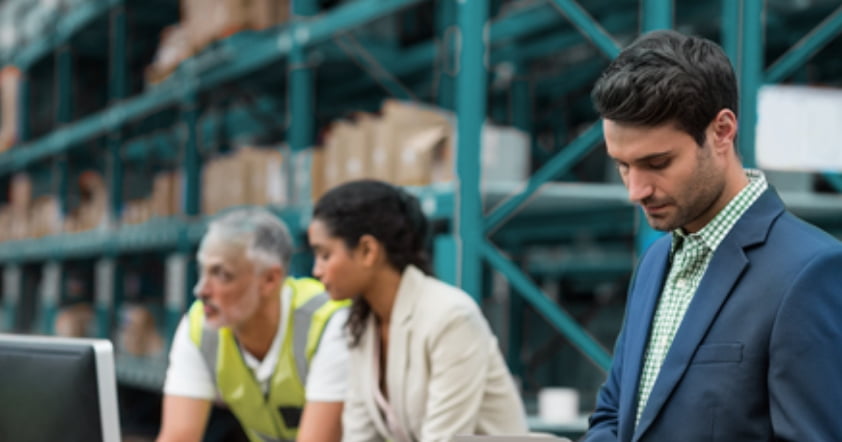  A man in a suit works on a laptop, while a woman and a man in a safety vest work on computers in a warehouse with shelves stacked with boxes, all connected through an advanced SIEM system for seamless managed detection and response.