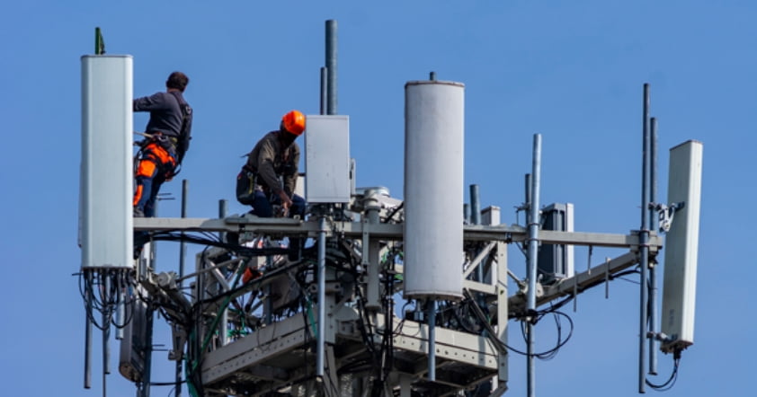 Two workers in safety gear are busy on a large telecommunications tower against a clear blue sky, implementing the latest technology to enhance Adaptive Managed xDR solutions.