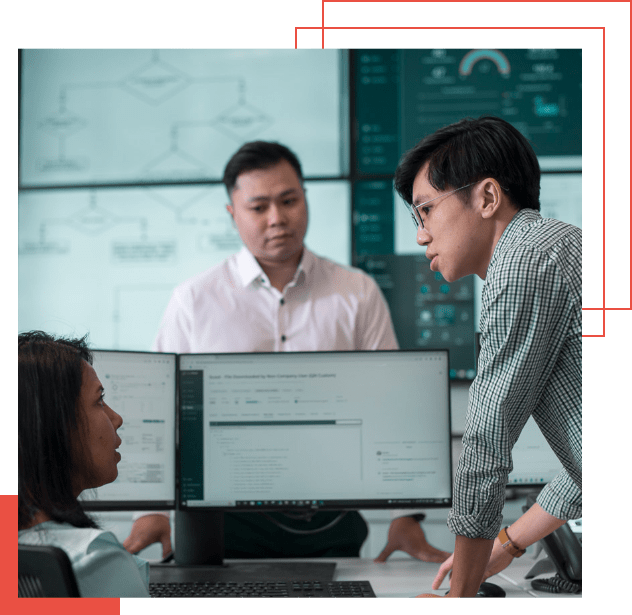 Drei Personen stehen und sitzen in einem Büro um einen Schreibtisch mit Computermonitoren herum und diskutieren über adaptive Managed xDR-Strategien. Im Hintergrund sind Workflow-Diagramme und ein digitales Dashboard zu sehen.