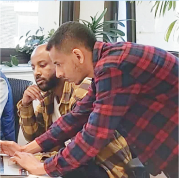  Dos hombres que visten camisas a cuadros trabajan juntos en un escritorio con una computadora portátil, discutiendo su última estrategia Adaptive Managed xDR en un entorno de oficina con plantas de fondo.