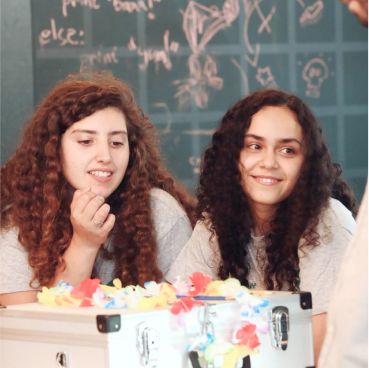  Two women with curly hair, wearing gray shirts, are smiling and sitting at a table with a decorated silver box. The chalkboard behind them is covered in drawings and text discussing Managed Detection and Response strategies.