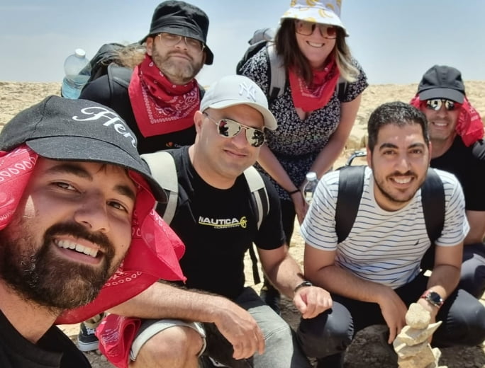  A group of six people, resembling an adventurous crew on a mission for Adaptive Managed xDR, poses together outdoors in a sunny, dry area with rocky terrain. They're all sporting hats, sunglasses, and bandanas.