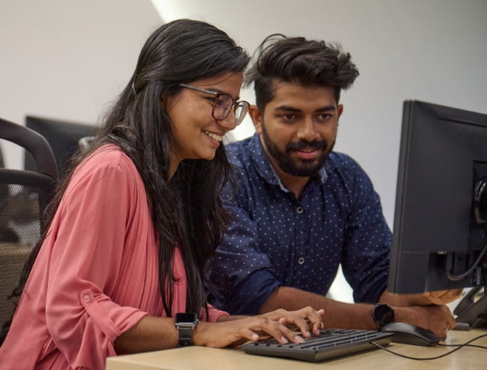  Two people are sitting in front of a computer monitor. One is typing on the keyboard while the other is looking at the screen, both smiling, as they delve into the intricacies of Managed Detection and Response (MDR).