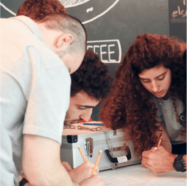 Three people work together, writing on papers placed on a table. They are using pencils and appear focused. A metal case, possibly containing notes on Adaptive Managed xDR, is also on the table.