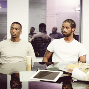  Two men sit side by side in a meeting room, engaged in conversation about Adaptive Managed xDR. A tablet, paper cup, and snacks are on the table in front of them. People are visible through a glass wall in the background.