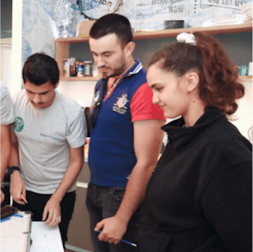 Three people are standing around a table, focused on an activity involving Managed Detection and Response. Two men are on the left and a woman on the right, with shelves holding various items in the background.