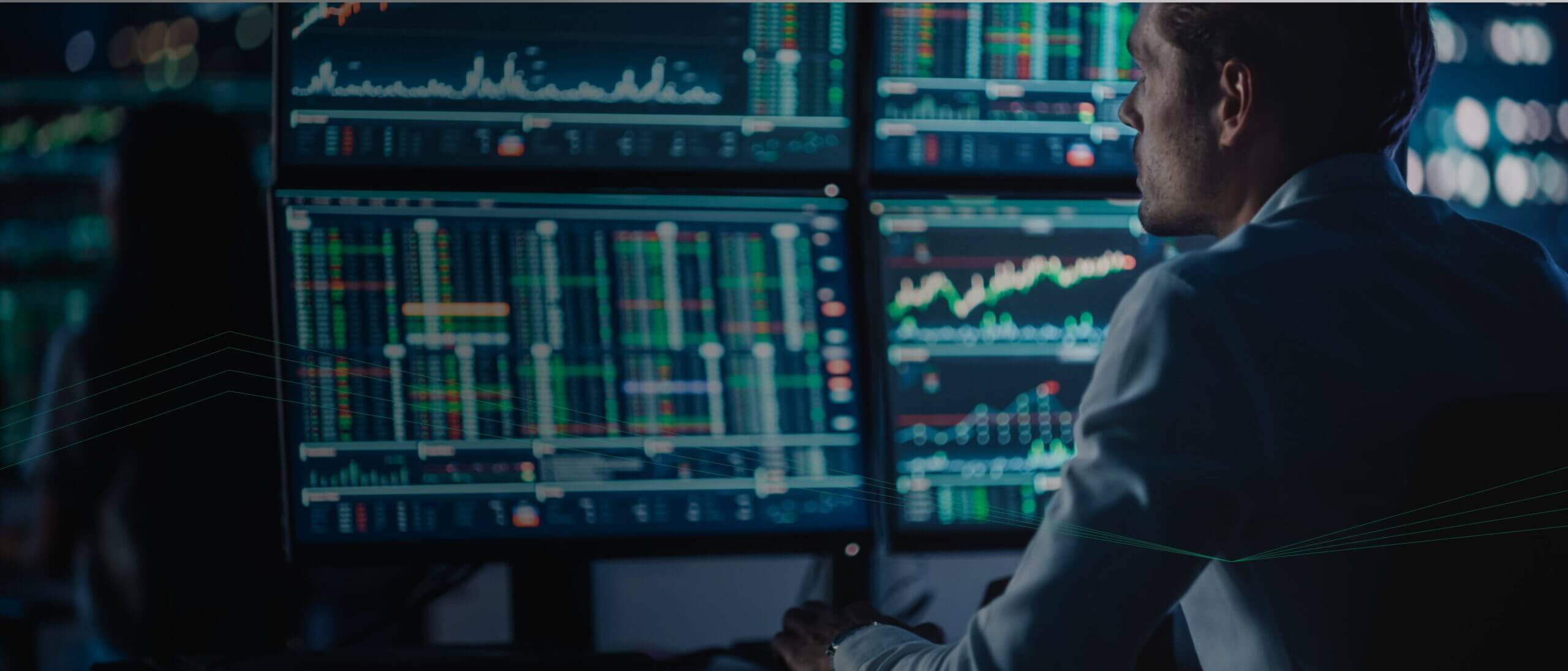 A person in a white shirt monitors multiple screens displaying financial charts and data in a dimly lit room, powered by the advanced analytics of Adaptive Managed xDR.