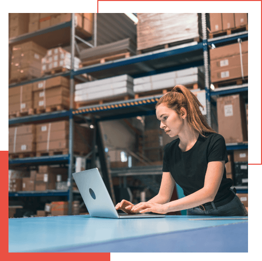  A person with a ponytail wearing a black t-shirt works on a laptop in a warehouse with shelves full of boxes in the background, focusing intensely on Managed Detection and Response solutions.