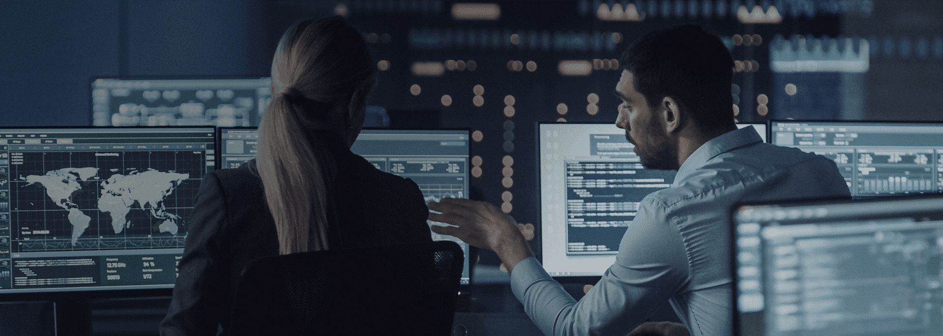 Two individuals sit in front of multiple computer screens, analyzing data and discussing information related to Managed Detection and Response in a dimly lit room with a cityscape visible through the window.