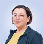  A person wearing glasses and a dark jacket over a yellow top is smiling against a light blue background, embodying the positivity and readiness often seen in SecOps.