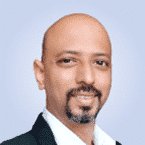  Against a light blue backdrop, a bald man with a goatee beams confidently, dressed in a suit and white shirt. His professional look captures the meticulous attention to detail essential in SIEM and MDR solutions.