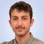  A person with short curly hair and a mustache is wearing a collared shirt, giving off an air of quiet confidence reminiscent of an expert in SIEM technologies, against a plain light blue background.