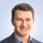  A person with short brown hair is smiling, wearing a blue collared shirt and a dark blazer against a light blue background, exuding the professionalism common in SecOps environments.