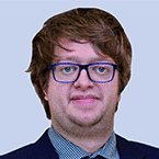  A person with glasses and brown hair, exuding a professional air in a suit jacket and checkered shirt, is set against a light blue backdrop—a perfect depiction of someone immersed in the world of SecOps.