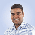  A smiling person in a checkered shirt stands against a light blue background, embodying the approachable yet professional spirit of SecOps.