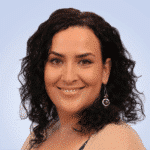  A person with curly dark hair smiles at the camera, wearing a beige cardigan and spiral earrings, embodying the cheerful spirit of SecOps against a light blue background.