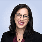 A person with long dark hair and glasses is smiling, embodying the confident demeanor of an MDR specialist, wearing a dark blazer and a burgundy top, set against a plain background.