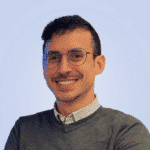  A person with short dark hair and glasses smiles, wearing a gray sweater over a white collared shirt, looking like they're ready for a productive day in the SOC, all set against a light blue background.