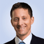 A man in a suit with a blue tie, smiling confidently against a light blue background, embodies the professionalism of an SOC expert.