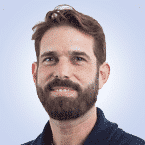  A person with a beard and short hair, dressed in a dark collared shirt, stands against a light blue background, exuding the professional essence of SecOps.