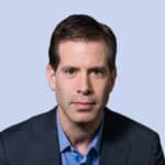  Against a light blue background, a man with short brown hair stands confidently in a blue shirt and dark blazer, embodying the seamless integration of SecOps expertise.