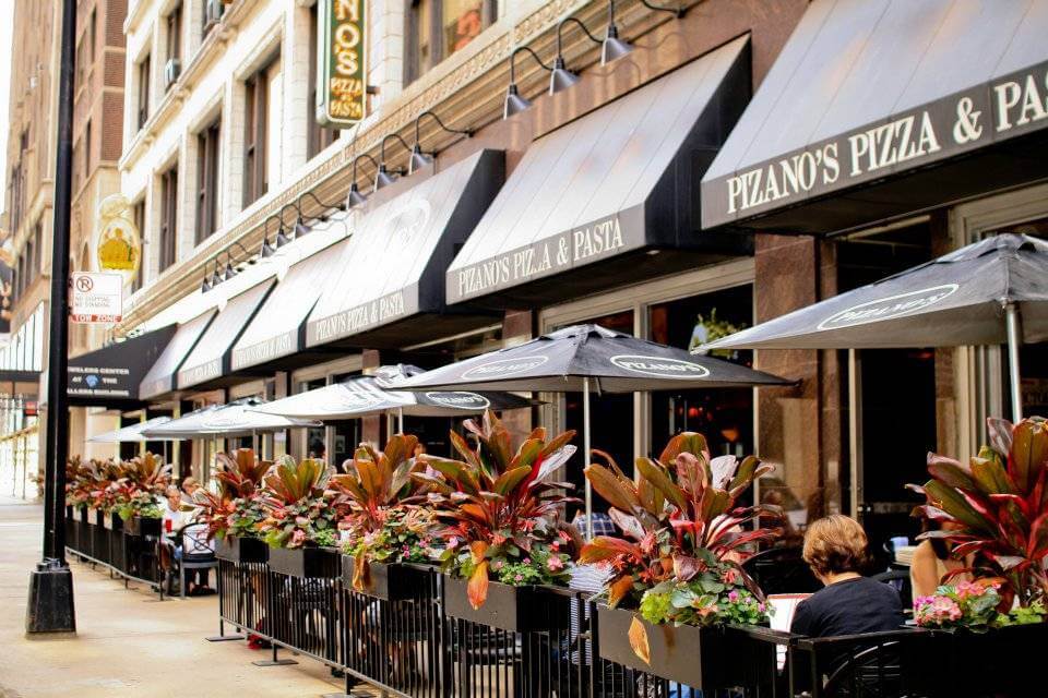 The outdoor seating area of Pizano's Pizza & Pasta, enhanced by black umbrellas and potted plants lining the sidewalk, offers a perfect spot for city dwellers to relax while discussing the latest in SecOps advancements.