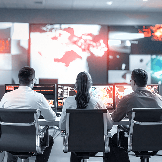 Three people sitting at desks monitor computer screens displaying global maps, data, and application security metrics, with a large map projection on the wall in a control room setting.