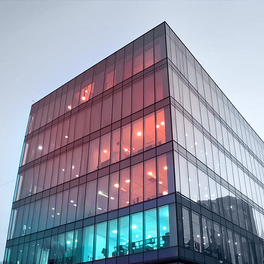 Glass office building with several floors, featuring colorful interior lighting in blue and red, photographed from a low angle against a clear sky—a modern space designed with cloud security in mind.