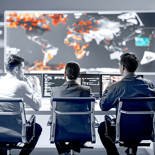 Three people sit at desks with computer screens, analyzing a large world map displaying red hotspots, suggesting global data monitoring or cloud security crisis tracking in a control room setting.