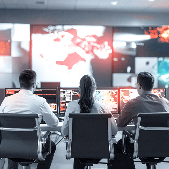 Three people sit at computer workstations in a control room, monitoring data and digital maps on large screens displaying global information and overseeing Identity & Access Management for secure operations.