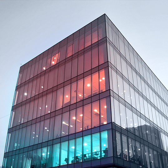 Modern glass office building with illuminated interior lights in red, blue, and white, viewed from a low angle against a clear sky—an ideal setting for advanced penetration testing to ensure robust digital security.