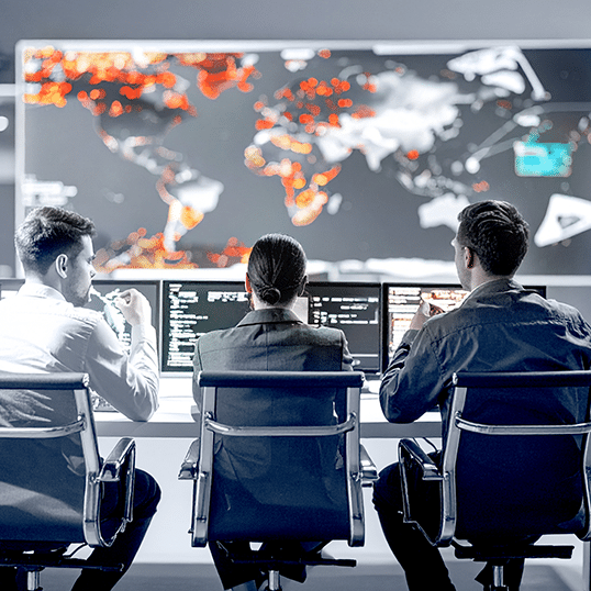 Three people sit at desks with computers, facing a large screen displaying a world map with highlighted red and orange areas, indicating global Penetration Testing results and potential vulnerabilities.