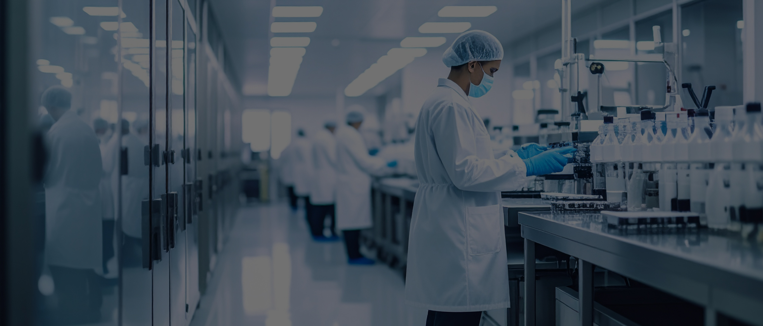 A laboratory technician in protective clothing works at a lab bench with bottles and equipment; others work in the background of this clean, well-lit healthcare laboratory.