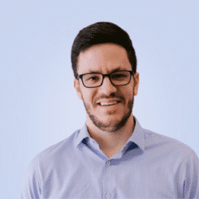  A man with short dark hair, glasses, and a beard wears a light blue button-up shirt and stands against a plain light background.