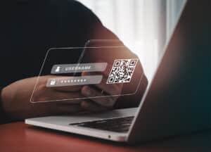 Person holding a smartphone in front of a laptop with a transparent overlay displaying username, password fields, and a QR code—illustrating the risks of QR code phishing and quishing attacks.