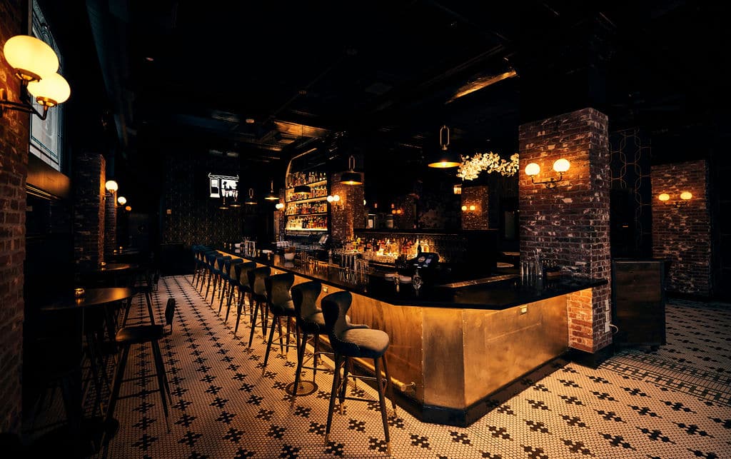 Dimly lit bar with brick walls, a brass counter, high-backed stools, and a patterned tile floor—perfect for a Lunch and Learn after a busy Microsoft Ignite 2025 conference; shelves of bottles and glasses stand ready behind the bar.