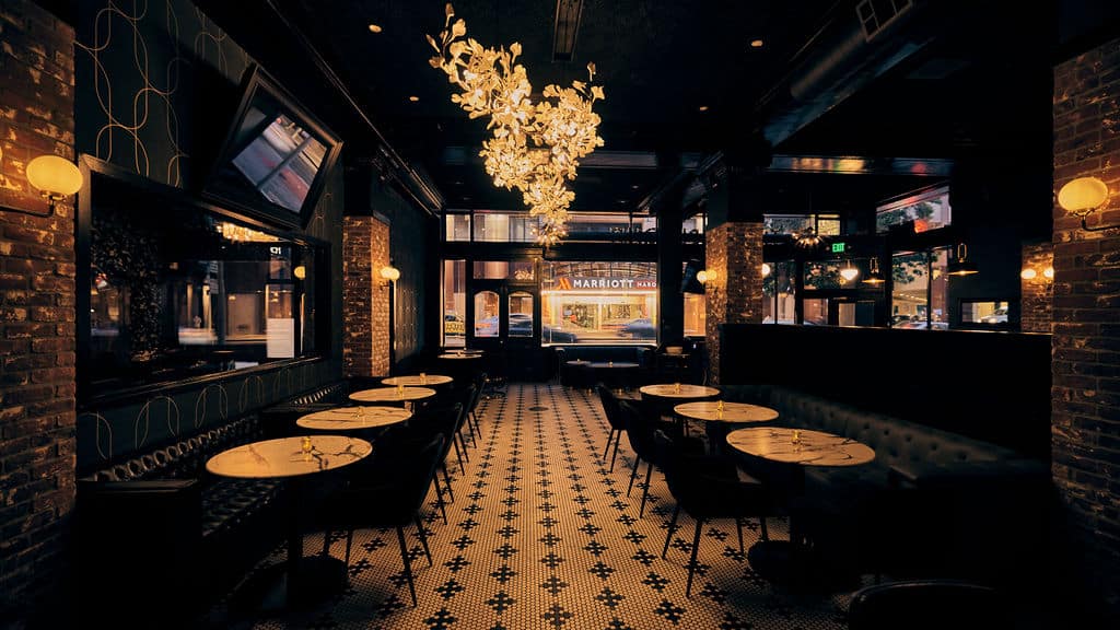 Dimly lit restaurant with marble tables, black chairs, patterned tile floor, exposed brick walls, and a decorative chandelier—perfect for a Lunch and Learn after Microsoft Ignite 2025; large windows face the street outside.