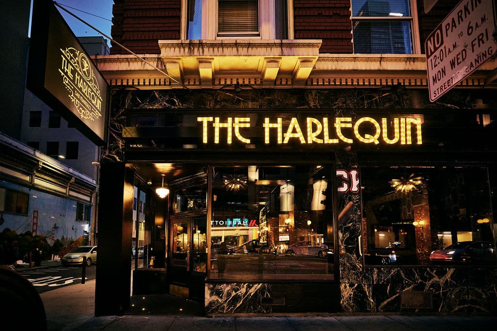 A street-level view of "The Harlequin" restaurant with illuminated signage and large front windows at night, perfect for a cozy Lunch and Learn or gathering after Microsoft Ignite 2025.