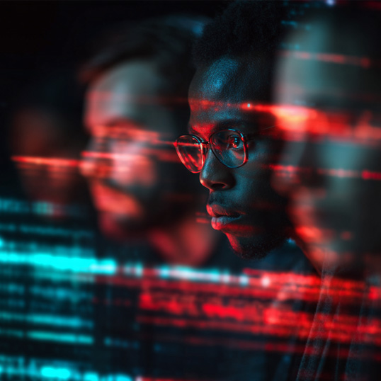  Three people stare intently at computer screens, illuminated by red and blue digital code overlays in a dimly lit room, reflecting the growing cybersecurity workforce and the challenges of addressing the security talent gap.