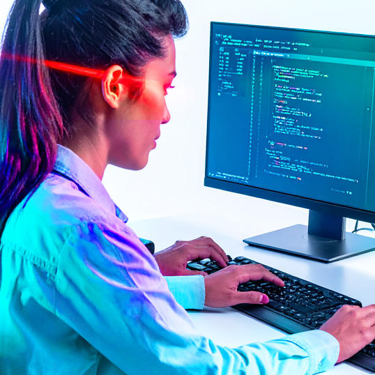  A woman in a blue shirt types code on a keyboard while looking at a monitor displaying programming scripts, highlighting her expertise amid the growing security skill shortage.