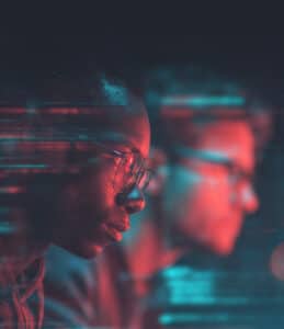 Two people in profile, illuminated by red and blue light, focus intently on computer screens with digital code effects overlaying the image.