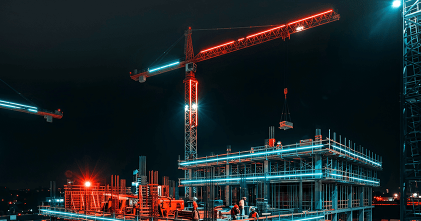 Construction site at night with workers, cranes, and an illuminated building framework highlighted by red and blue lights.
