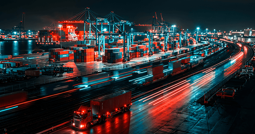 Shipping containers stacked at a busy port at night, with cranes, trucks, and blurred lights from moving vehicles creating a vibrant scene.